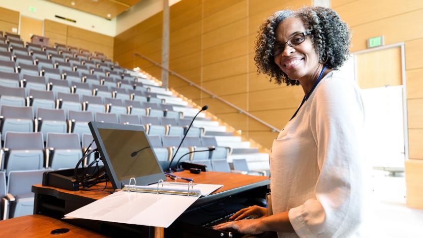 Smiling, older, female professor stands at lectern with laptop, binder and microphone, in front of large classroom of empty theater seating.