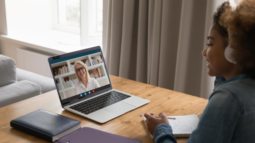 Smiling young woman in jean jacket wearing white headphones sits at wooden desk holding pencil with notebook, looking into laptop at a video call of a smiling, older, blond woman in white shirt who sits in front of bookshelves, seen on screen of computer.