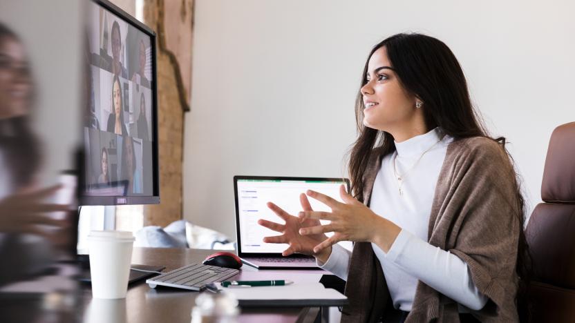 Woman sits at desk, talking animatedly with her hands, looking into computer monitor of many people on screen. Laptop open beside her on the desk next to coffee cup.