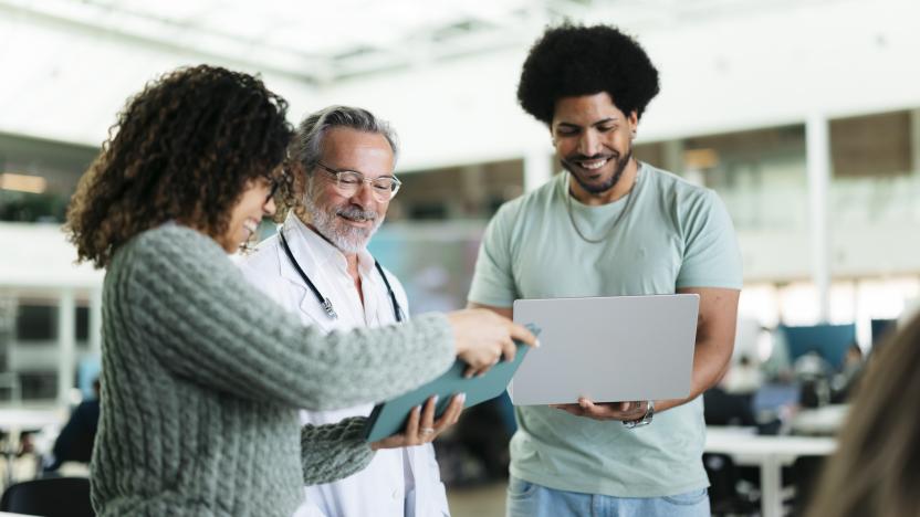 Smiling young man and woman helping older instructor man in white lab coat wearing stethoscope with laptop and tablet.
