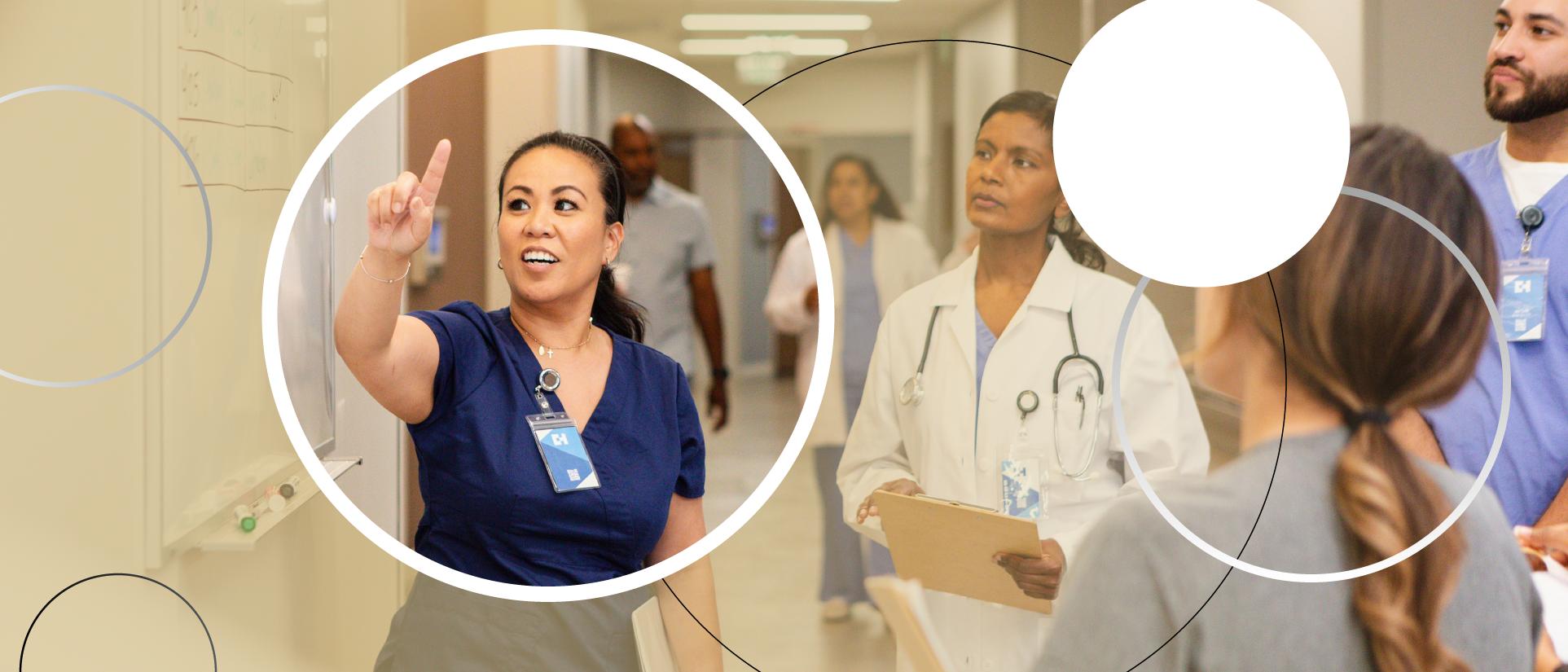 Dark-haired nurse woman in scrubs points at a white board in a hospital breezeway, surrounded by a doctor woman in white lab coat with stethoscope around her neck, another nurse man in scrubs and woman in grey t-shirt look on intently. More medical staff walk toward the group from down the hall. 