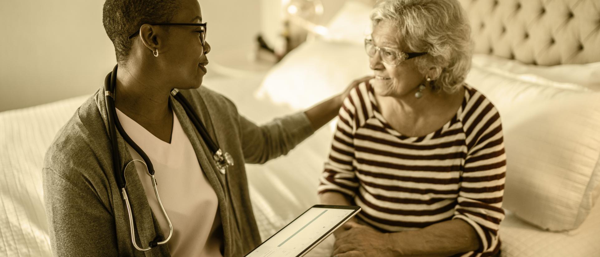 Nurse with a stethoscope around her neck puts a caring arm on the shoulder of an elderly woman as they sit on a bed in a home.