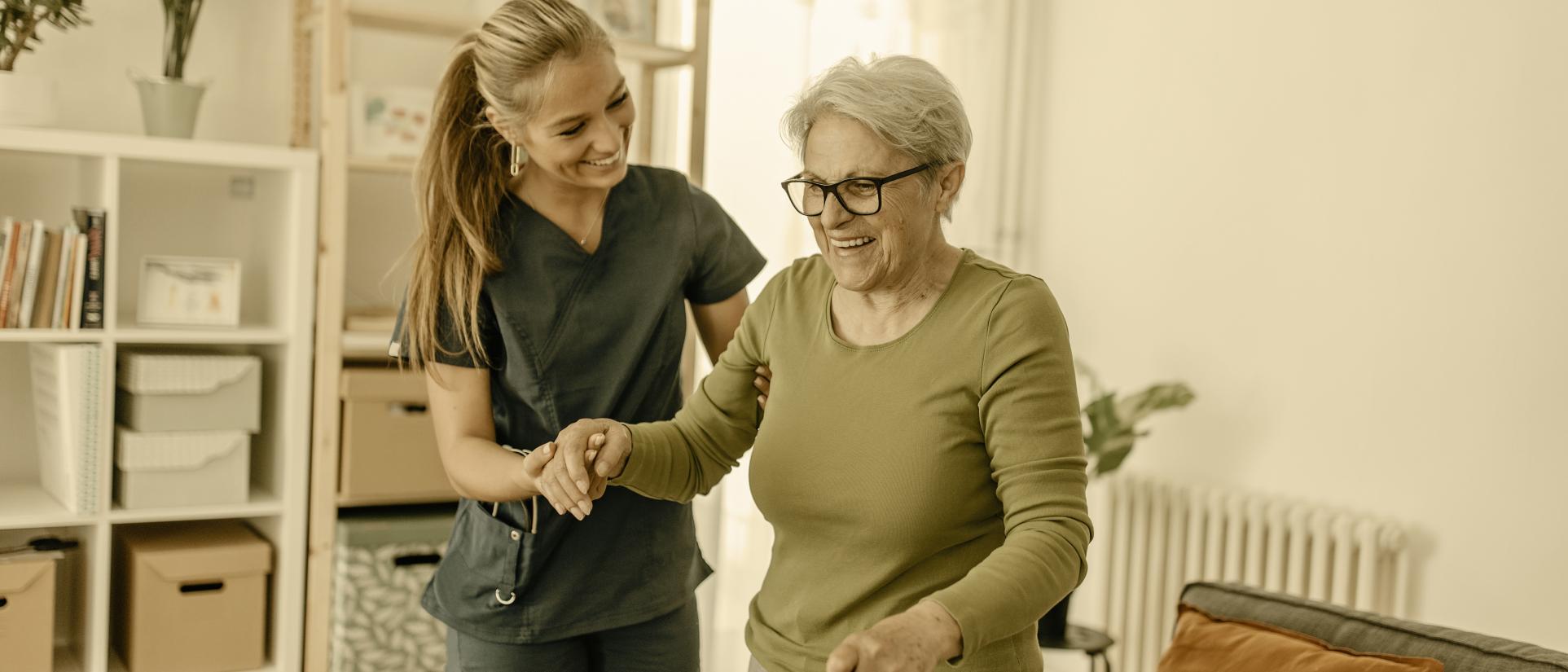 An older woman walking with a cane receives assistance from a woman in scrubs.
