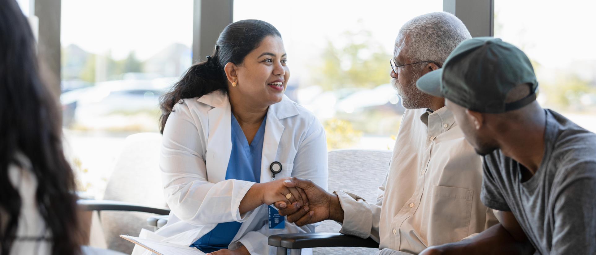 A woman wearing a hospital badge and a white coat holds the hand of a patient in plain clothes while they talk and a family member looks on.