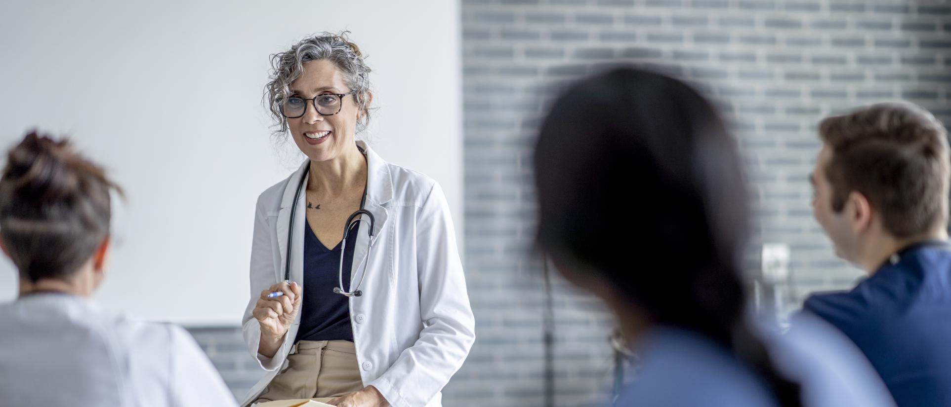 Older woman with curly grey hair wearing a white doctor's coat and stethoscope sits in front of 3 nursing students in scrubs in a teaching setting.