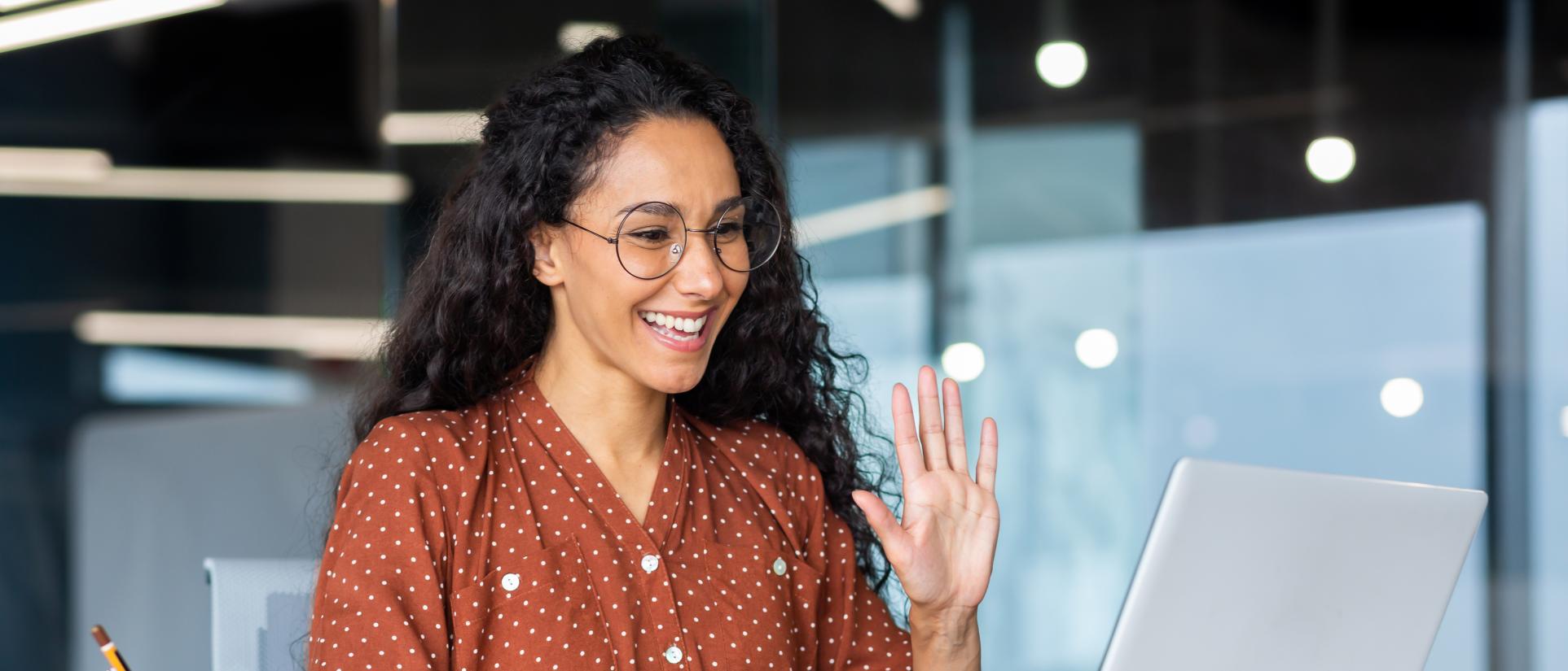 Smiling young woman in brown, polka-dot blouse waves at laptop computer on desk in front of her.