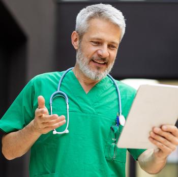 Smiling grey haired man in green scrubs with stethoscope, holding a tablet.