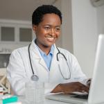 Smiling woman with short curly hair, white lab coat, stethoscope, types on laptop computer sitting at table at home.