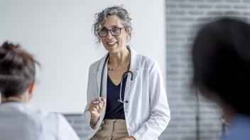 Older woman with curly grey hair wearing a white doctor's coat and stethoscope sits in front of nursing students in a teaching setting.